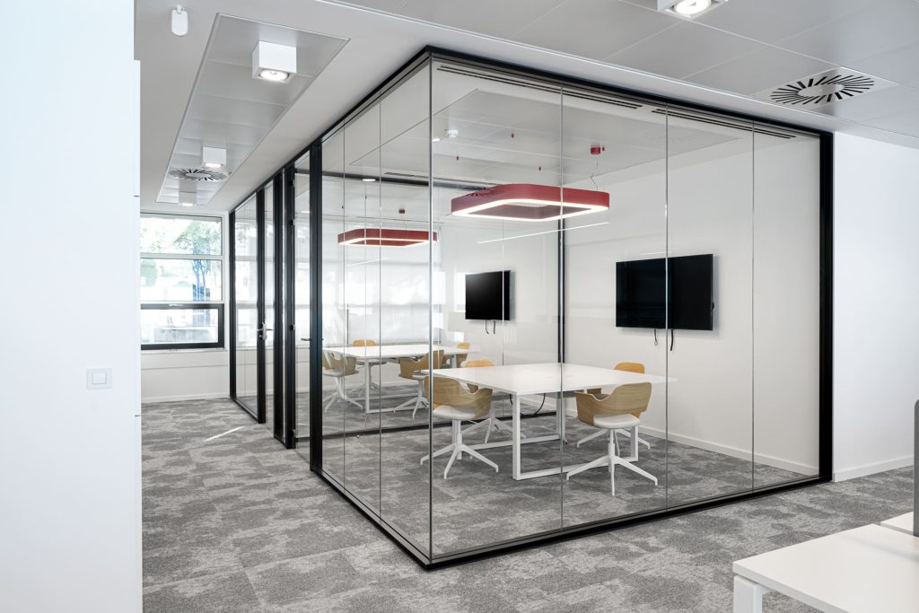 Oficina moderna con una sala de reuniones acristalada, delimitada por paneles de vidrio con marcos negros. En el interior hay una mesa blanca rectangular rodeada por sillas de madera clara con base blanca. Dos lámparas colgantes con estructura cuadrada roja y luz blanca se ubican sobre la mesa, mientras que dos pantallas planas están montadas en las paredes opuestas. El espacio exterior está cubierto con una alfombra gris y tiene paredes blancas, lo que refuerza la sensación de limpieza y amplitud.