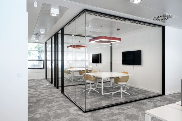 Oficina moderna con una sala de reuniones acristalada, delimitada por paneles de vidrio con marcos negros. En el interior hay una mesa blanca rectangular rodeada por sillas de madera clara con base blanca. Dos lámparas colgantes con estructura cuadrada roja y luz blanca se ubican sobre la mesa, mientras que dos pantallas planas están montadas en las paredes opuestas. El espacio exterior está cubierto con una alfombra gris y tiene paredes blancas, lo que refuerza la sensación de limpieza y amplitud.