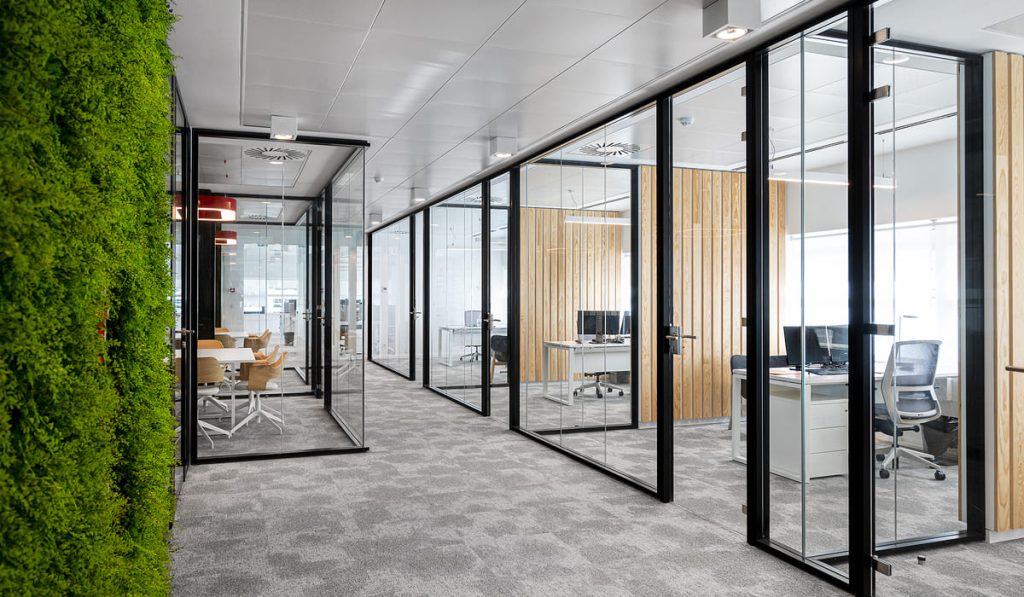 Pasillo de oficinas modernas con divisiones de vidrio y marcos metálicos negros, que separan distintas salas de trabajo. A la izquierda, una pared verde con vegetación natural o artificial aporta un toque de frescura y sostenibilidad al ambiente. En las oficinas se observan escritorios blancos con sillas ergonómicas, ordenadores y paredes de madera clara que contrastan con el diseño industrial del vidrio y el metal. El suelo está cubierto por una alfombra gris que recorre todo el espacio, mientras que las luces empotradas y lámparas colgantes proporcionan una iluminación cálida y equilibrada.